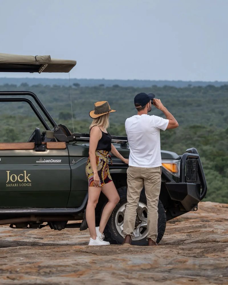 Two guests beside a Jock Safari Lodge game vehicle on a rocky outcrop above the Kruger