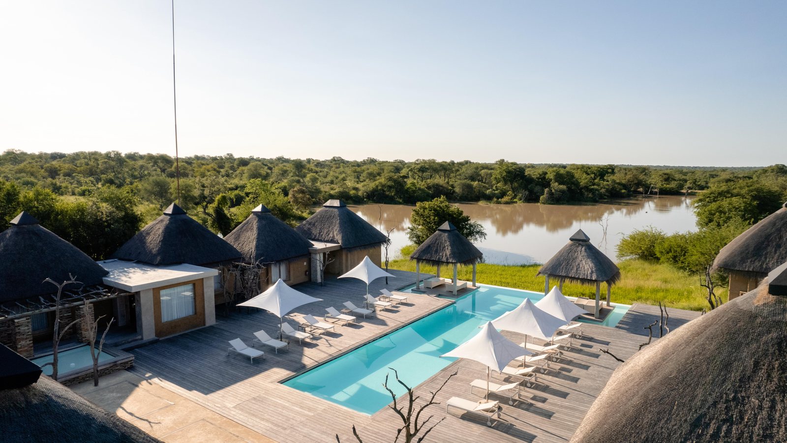 Kapama Karula Lodge seen across the Klaserie River, its thatched suites and pool deck set against the bush of the Kapama Private Game Reserve