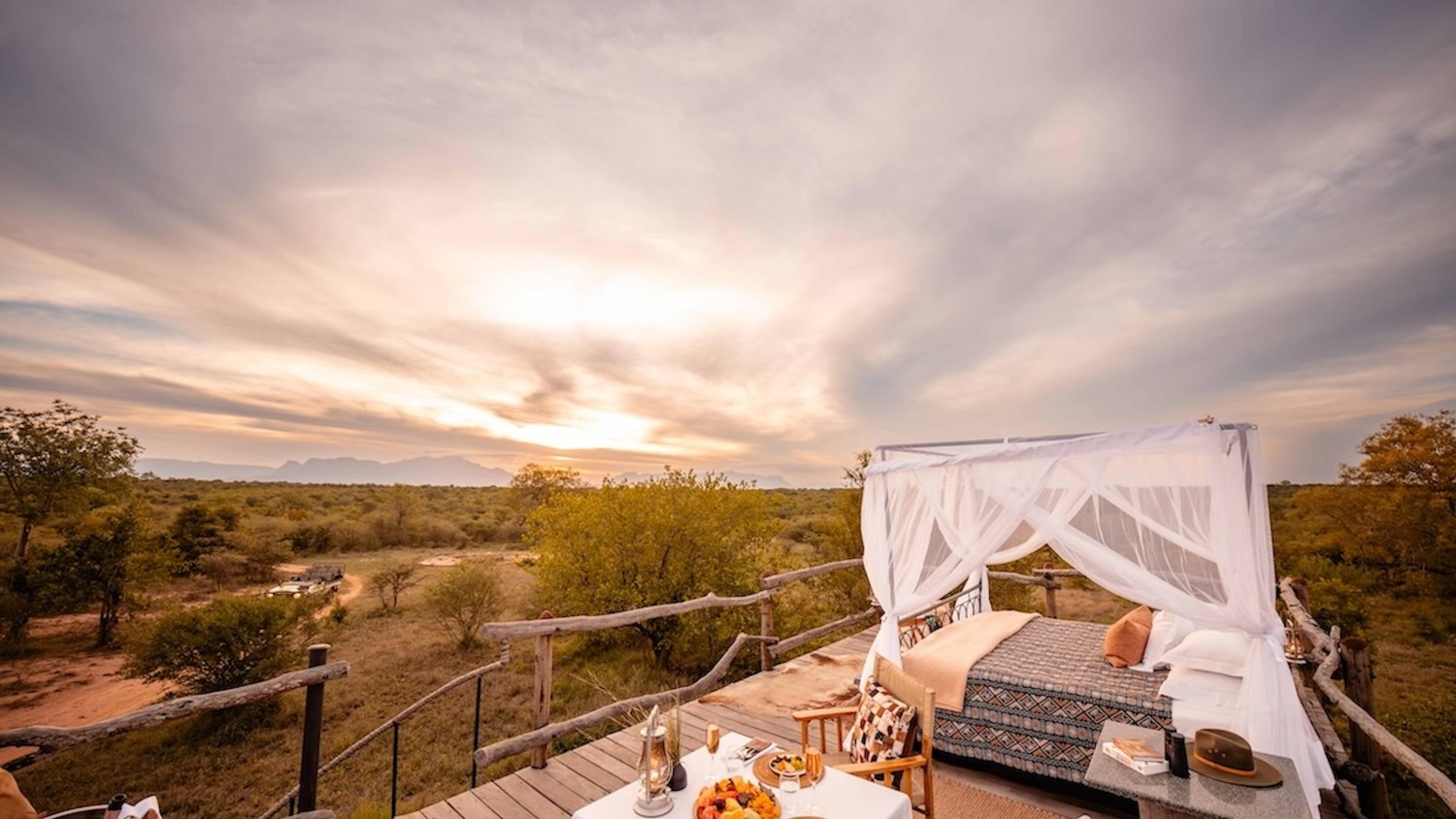 A four-poster bed under a mosquito net on Kapama's open-air sleep-out platform, set against a sunset over the reserve